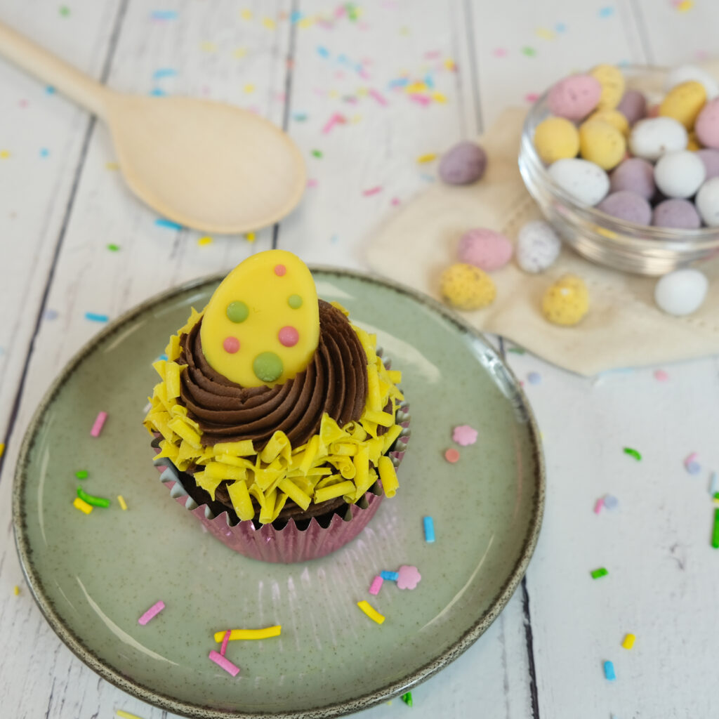 Easter themed cupcake with yellow icing decor and an icing easter egg on top, sitting on a green plate on a table with colourful sprinkles scattered around. Mini eggs in a dish and a wooden spoon in the background.